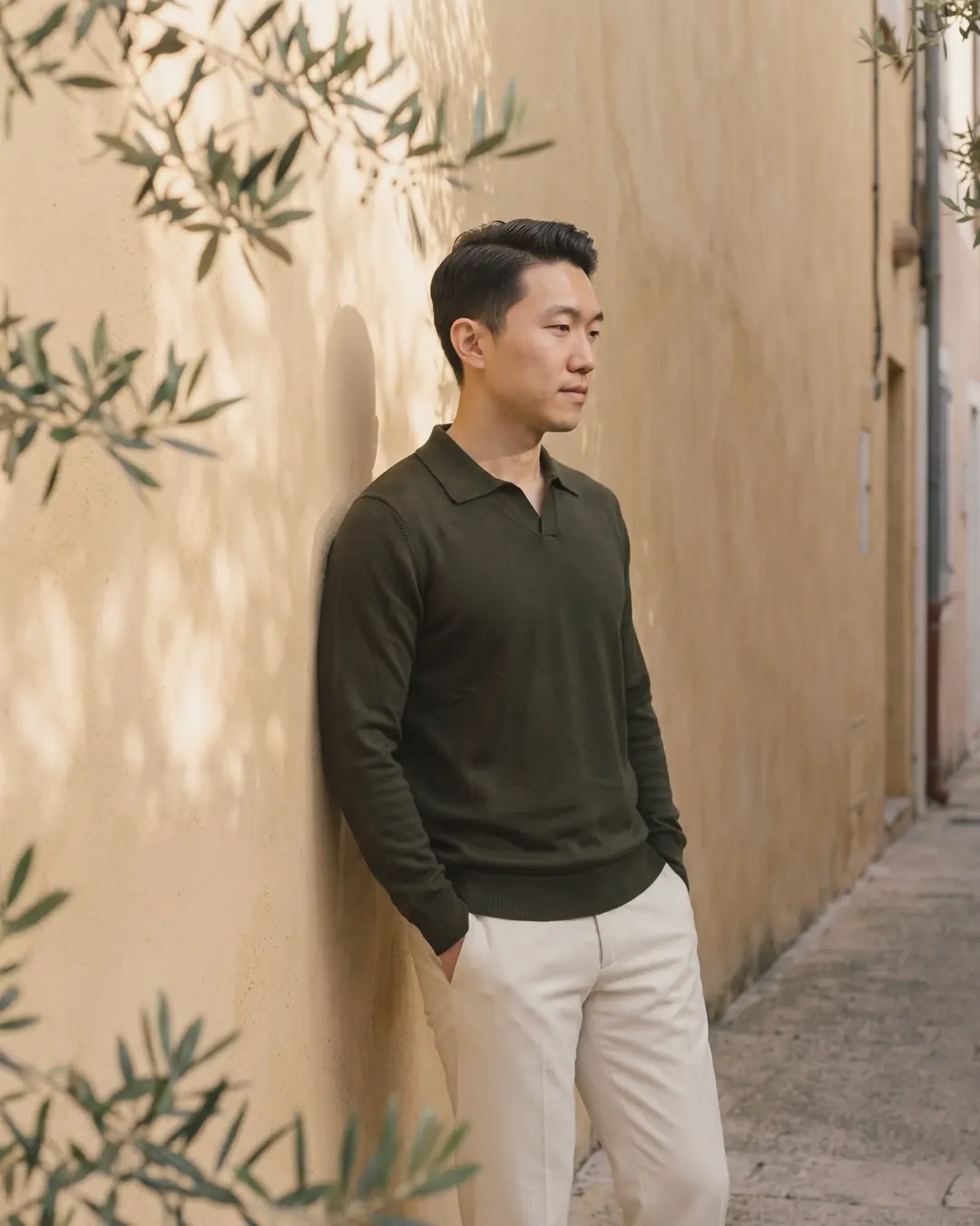 A man in an olive knit polo and ecru trousers leaning on a sunlit ochre wall