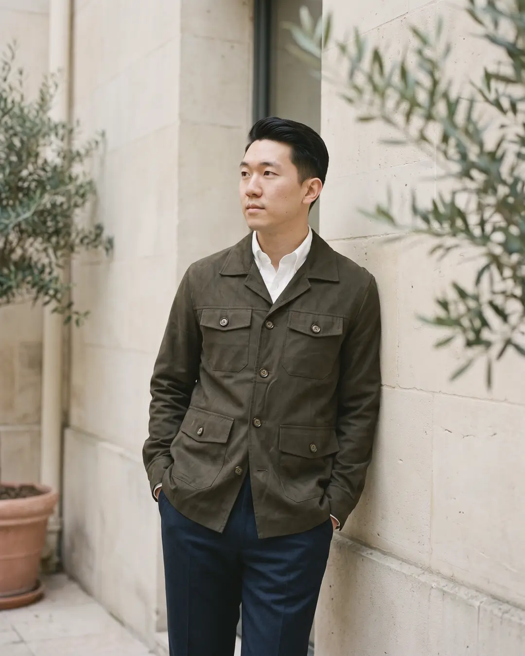 A man in an olive overshirt over a white Oxford shirt and navy trousers against a stone courtyard wall